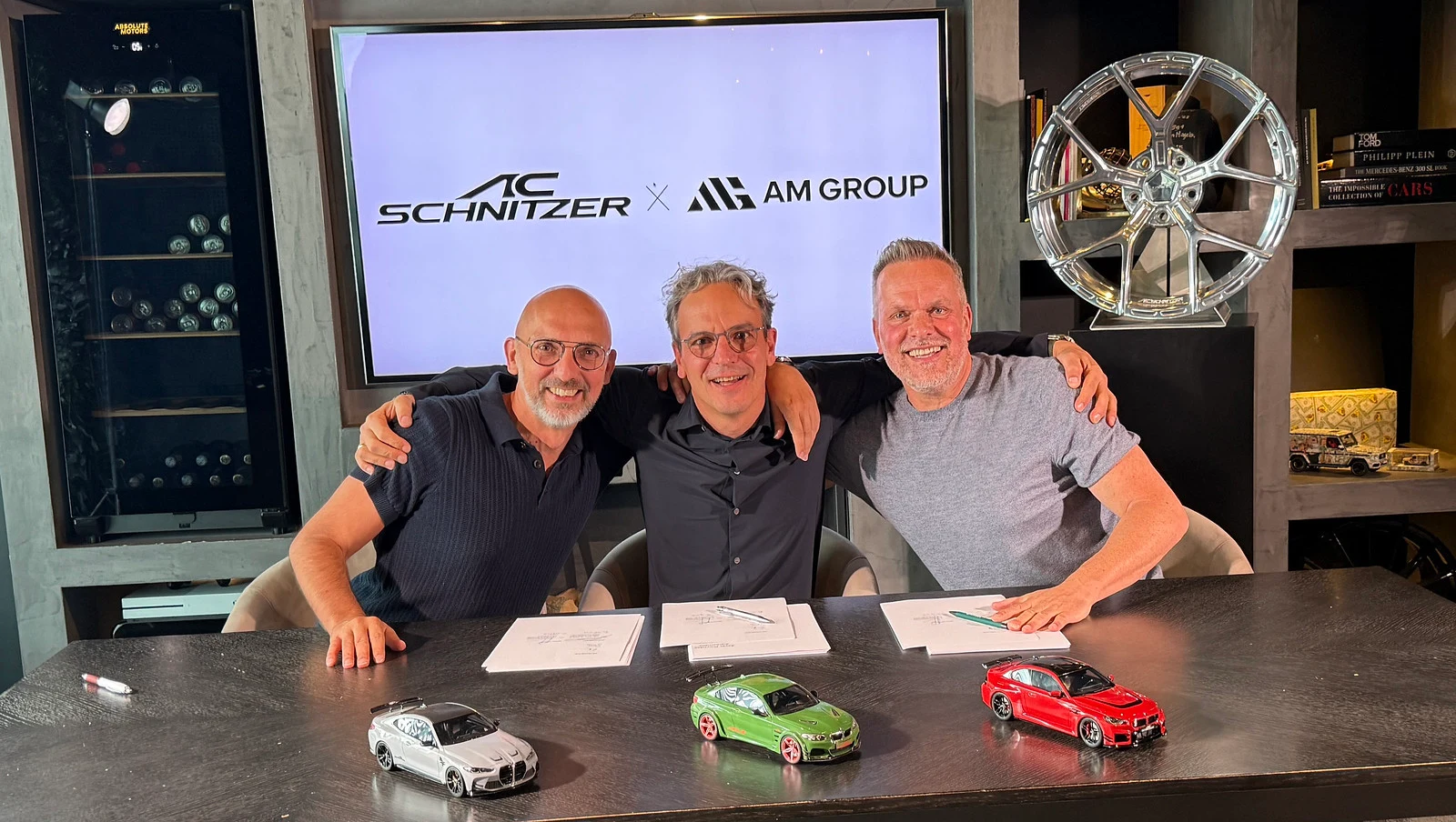 AC Schnitzer and AM Group representatives at the transfer announcement, seated in front of an AC Schnitzer × AM Group sign with three miniature BMW models and a displayed alloy wheel.